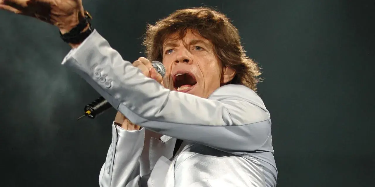 Mick Jagger, wokalista zespołu, śpiewa z mikrofonem, gestykulując ręką.