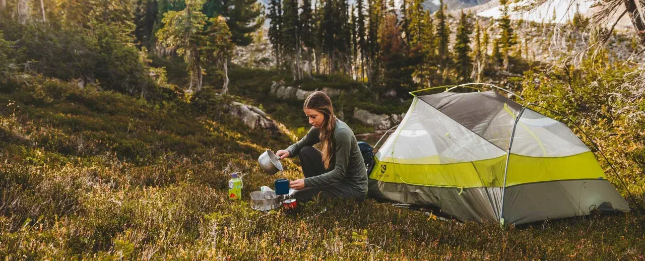 A woman enjoys a peaceful moment of camping alone, preparing a warm drink next to her tent amidst a serene forest landscape.