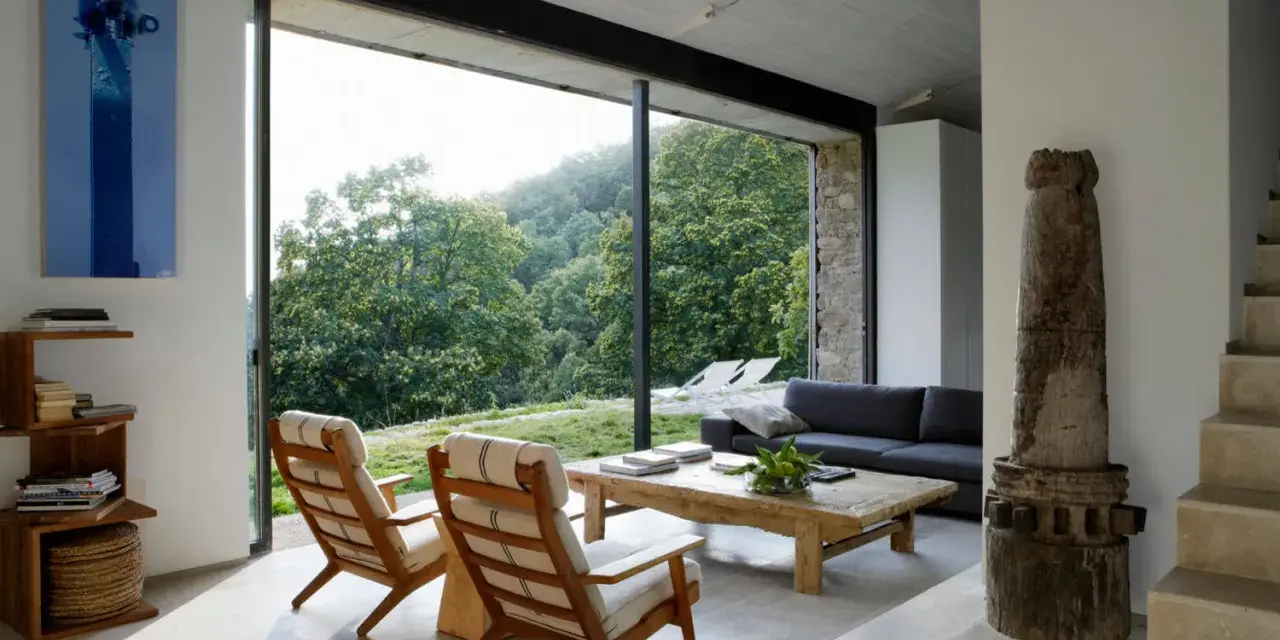 Salón moderno con sofás, sillones y mesa de centro rústica. Los suelos de piedra, con un estilo que evoca los años 70, contrastan con la vista exterior de un bosque frondoso.