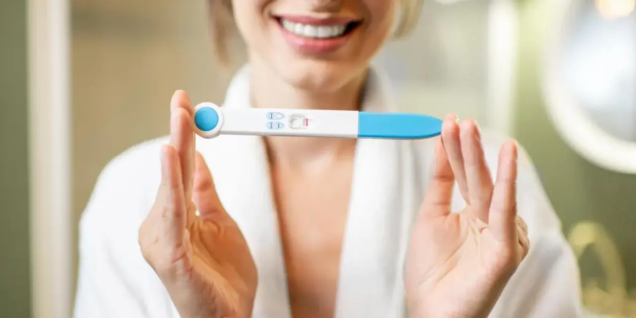 Uśmiechnięta kobieta w szlafroku trzyma test ciążowy z dwoma kreskami. To moment, kiedy warto zastanowić się, kiedy zrobić test ciążowy.