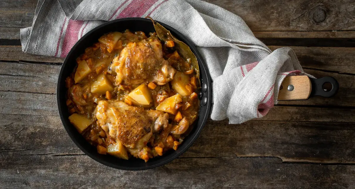 Estofado de pollo con patatas y zanahorias en sartén sobre mesa de madera.