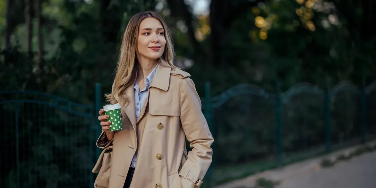 Donna con trench beige, un classico del marchio Burberry, sorseggia un caffè all'aperto.
