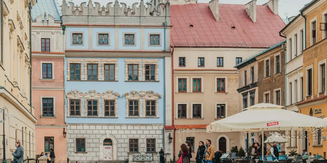 Kolorowe kamienice na rynku, idealne na tani city break. Ludzie spacerują, siedzą w ogródku kawiarnianym.