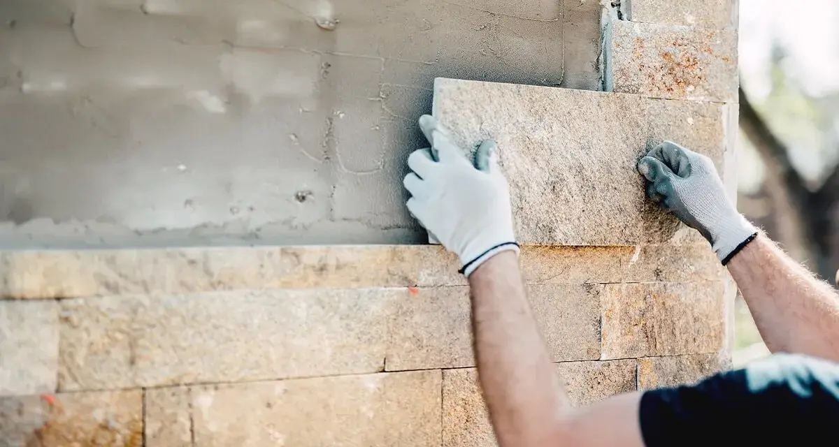 Ręce w białych rękawiczkach precyzyjnie wykonują układanie kamienia elewacyjnego na zewnątrz. Powstaje piękna, kamienna ściana.