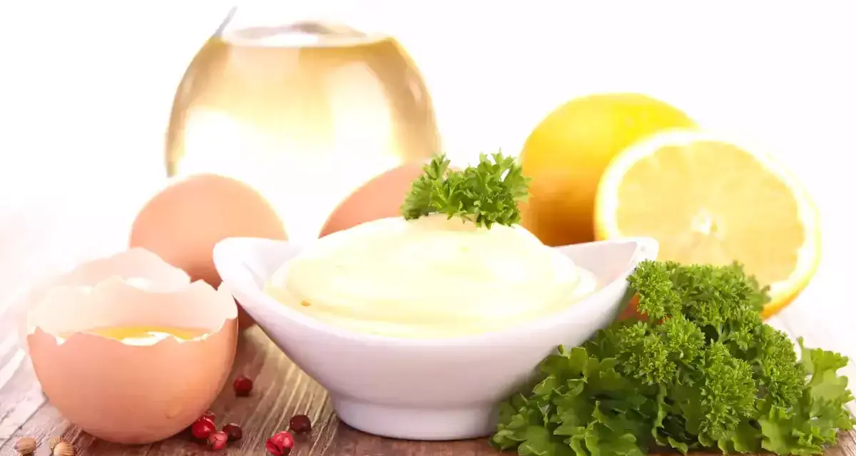 Un bol de mayonnaise maison crémeuse, entouré d'œufs, de citron, d'huile et de persil.