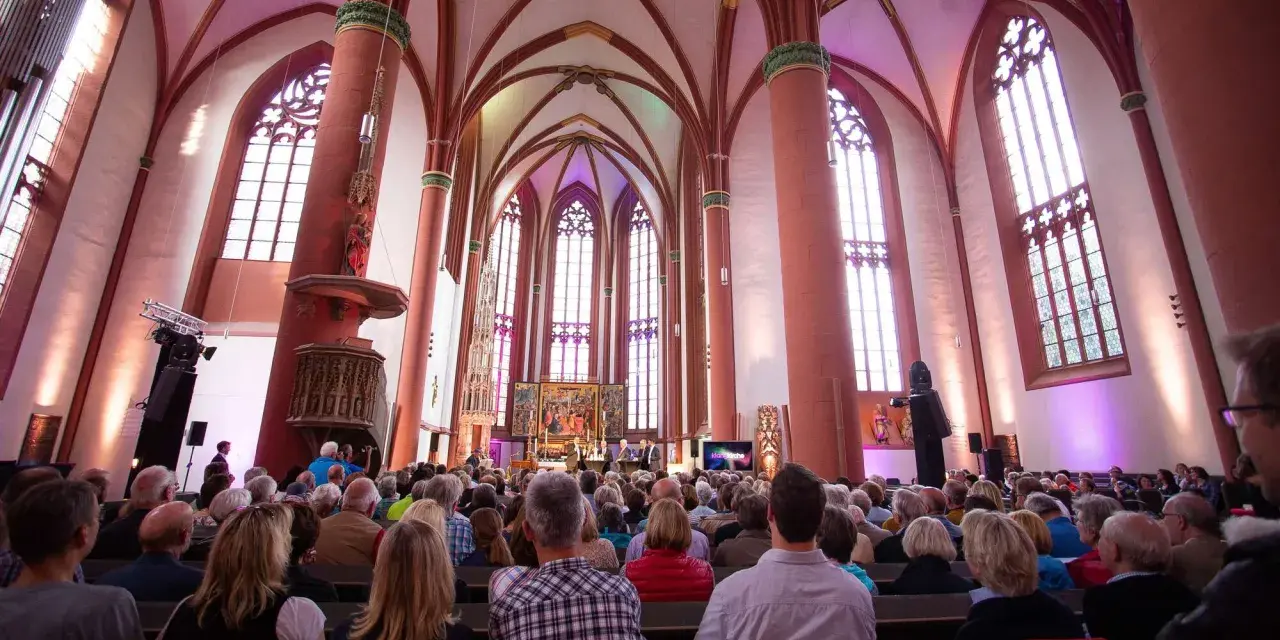 Menschen sitzen in einer Kirche, die für eine Veranstaltung beleuchtet ist. Es ist schön, dass man wieder in die Kirche eintreten kann.