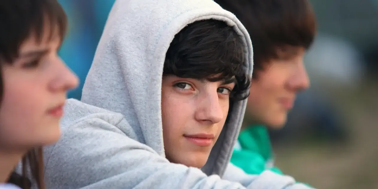 Drei Jungs, etwa 13 Jahre alt, sitzen nebeneinander. Einer trägt einen grauen Kapuzenpullover, der sein Gesicht fast verdeckt.