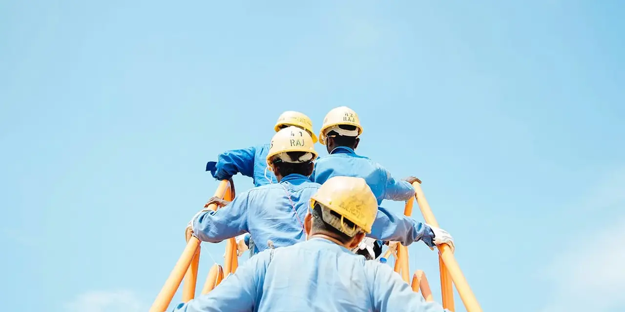 Trabajadores con cascos amarillos suben una escalera naranja contra un cielo azul claro. Podría ser una escena de cesión ilegal de trabajadores.