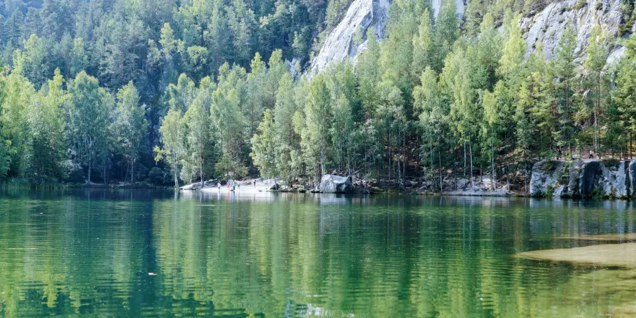 Zielona tafla jeziora odbija las i skalne miasto w Polsce. Ludzie spacerują po piaszczystym brzegu.