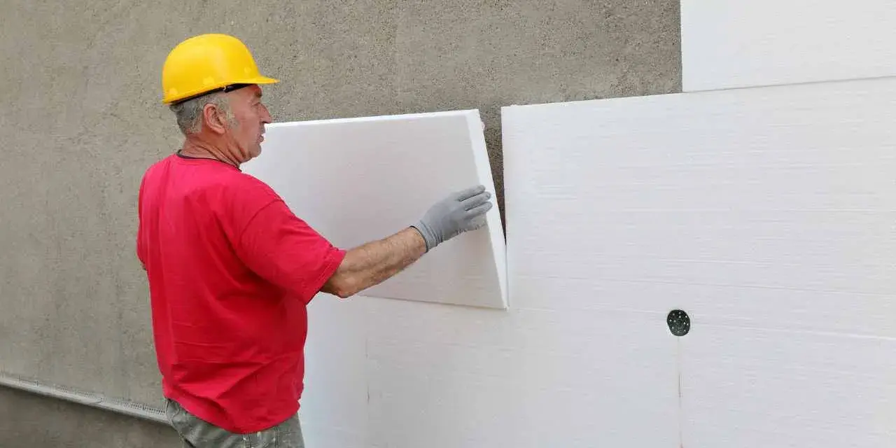 Operaio con casco giallo applica pannelli isolanti bianchi. Attenzione: il polistirolo è infiammabile.