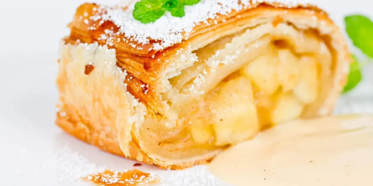 Ein Stück Apfelstrudel mit Filoteig, bestäubt mit Puderzucker und garniert mit Minze, serviert mit Vanillesauce.