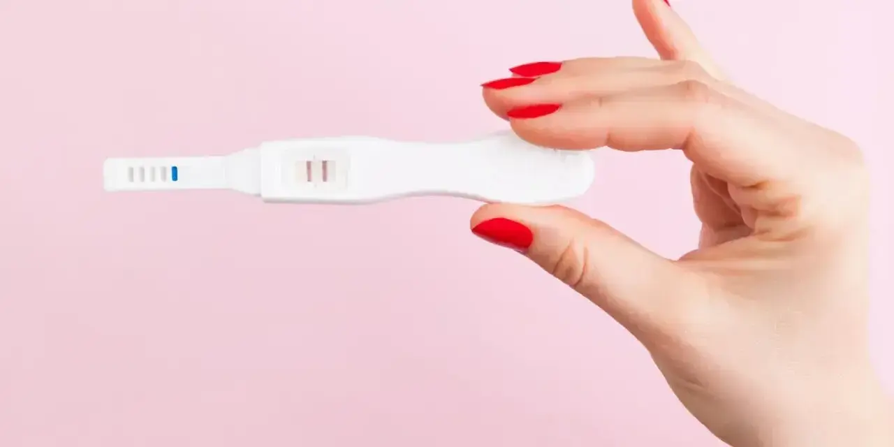 Dłoń z czerwonymi paznokciami trzyma test ciążowy z dwoma kreskami. To może być sygnał, że doszło do ciąży biochemicznej.