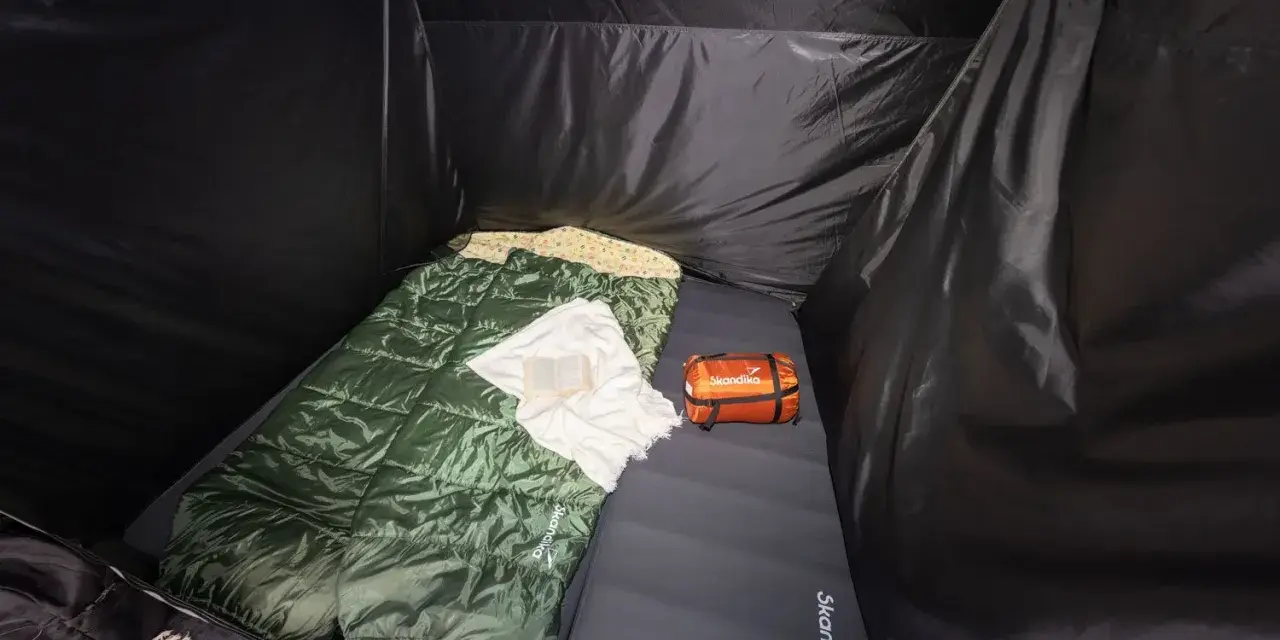 Schlafsack und Kissen auf der Isomatte, welche Seite oben ist, ist klar erkennbar. Ein kleines orangefarbenes Päckchen liegt daneben.