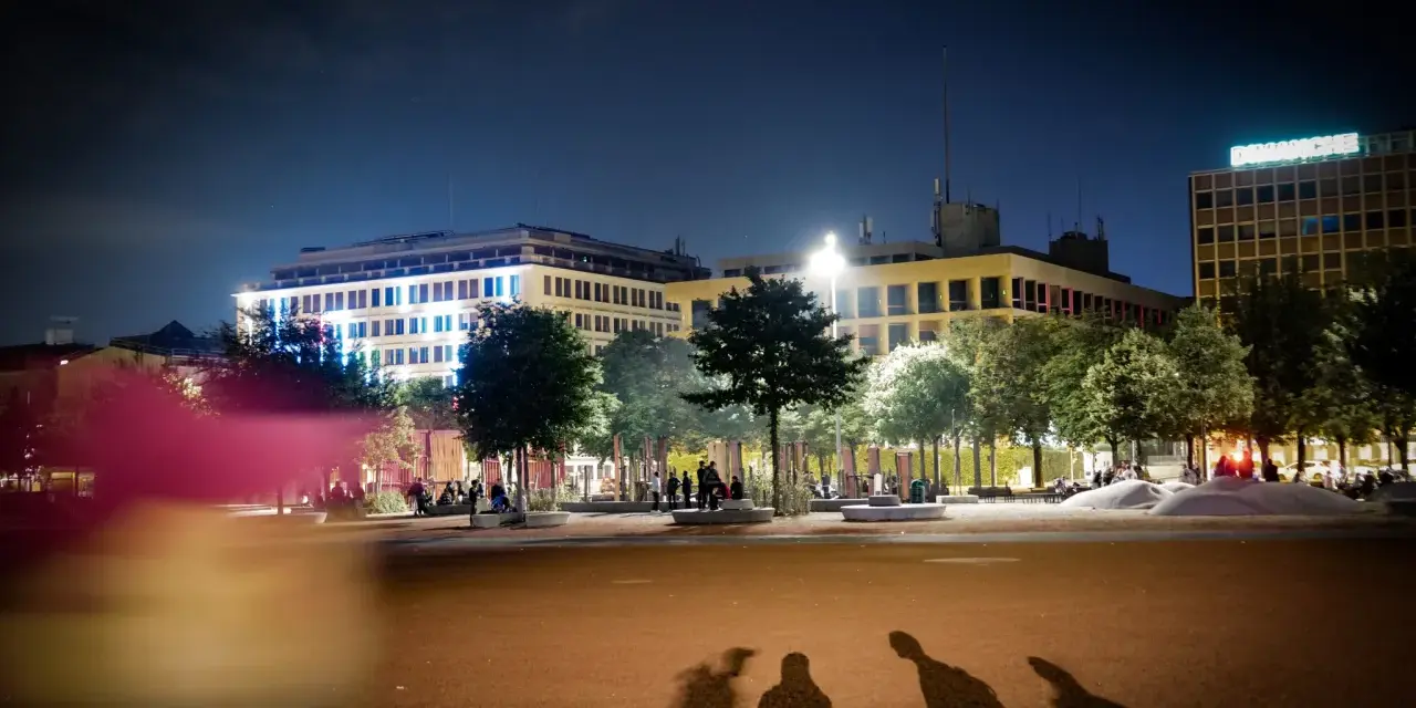 Ombre d'un mineur dehors la nuit, projetées sur une place éclairée par des bâtiments modernes.