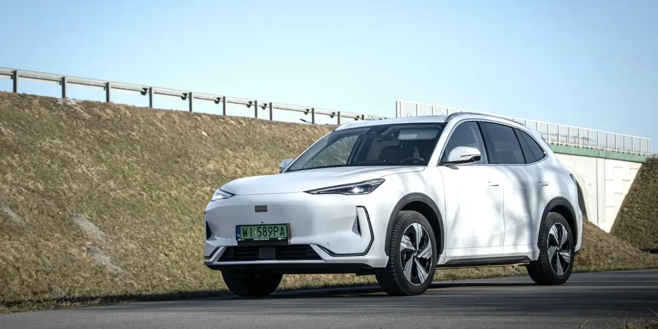 Biały SUV Geely E5 (EX5) na tle wiaduktu i niebieskiego nieba.