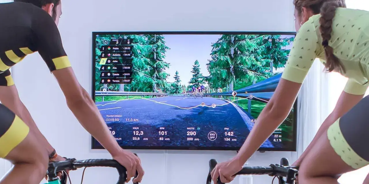Two cyclists race virtually on a scenic road using indoor cycling apps. The screen displays their stats and rankings.