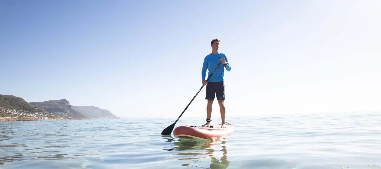 Mann steht auf SUP-Board, hält Paddel richtig. Ruhige See, bergige Küste im Hintergrund.
