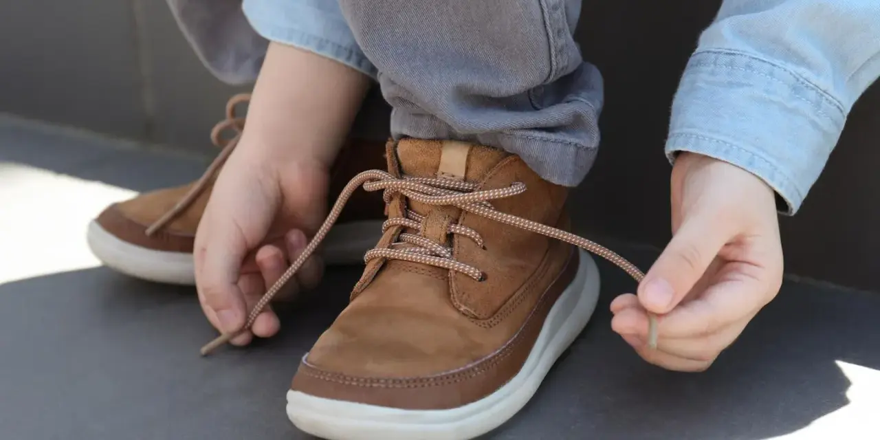 Kind lernt, seine braunen Schnürstiefel zu binden. Die Schnürsenkel sind hellbraun und gemustert.