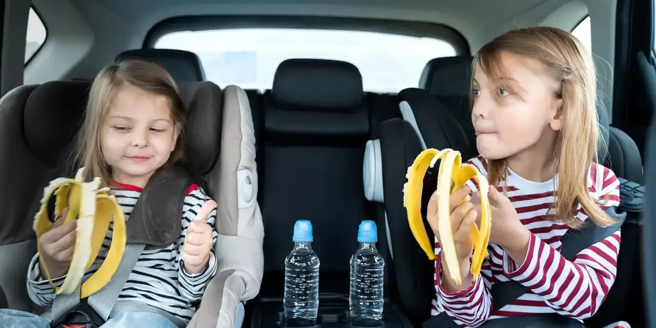 Dwie dziewczynki w fotelikach samochodowych jedzą banany. Woda i przekąski to podstawa, gdy zastanawiasz się, co zabrać na długą podróż samochodem z dzieckiem.