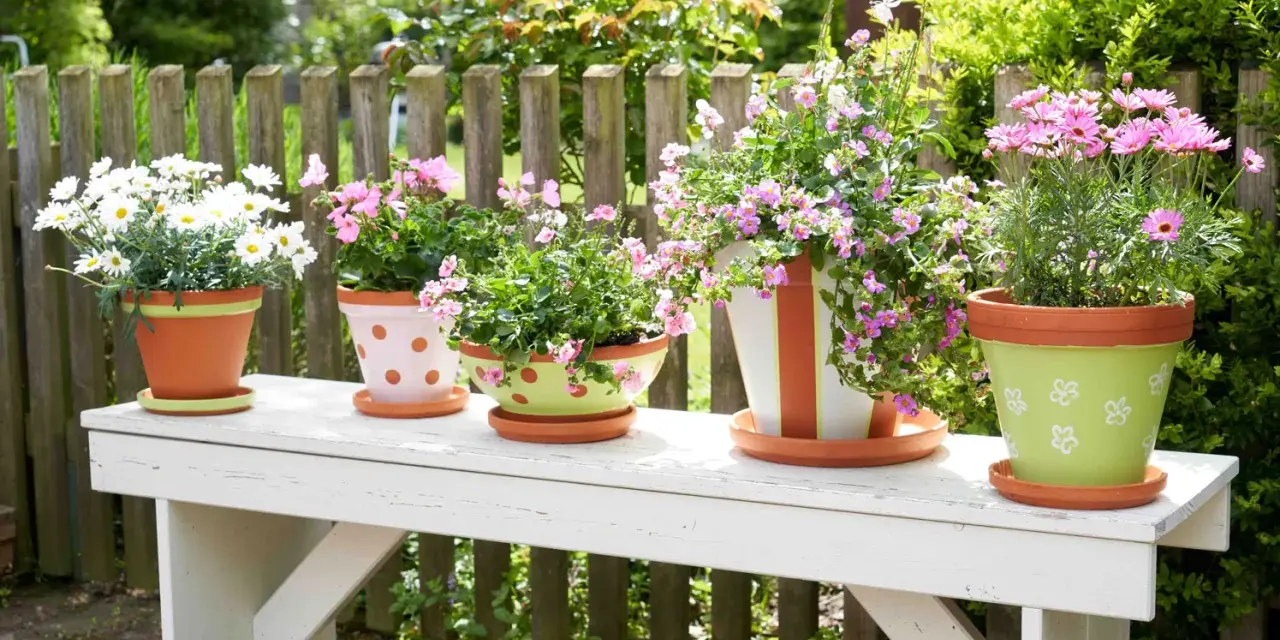 Fünf bemalte Blumentöpfe mit bunten Blumen stehen auf einer weißen Bank im Garten. Ein Blumentopf ist grün mit weißen Blumen bemalt.