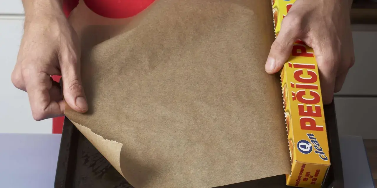 Hände legen Backpapier auf ein Blech. Die Rolle mit der Aufschrift "PEČÍCÍ PAPÍR" zeigt, dass man Backpapier einfetten muss, um ein Anhaften zu verhindern.