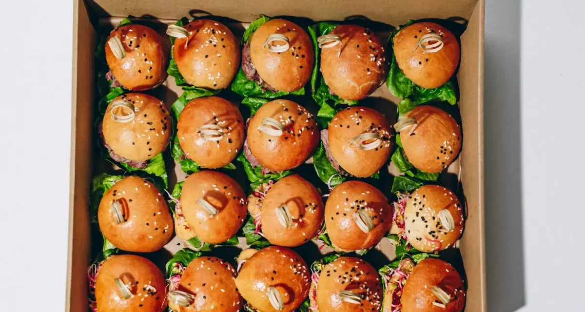 Pudełko z dwudziestoma mini burgerami na liściach sałaty, posypane sezamem i ozdobione patyczkami.
