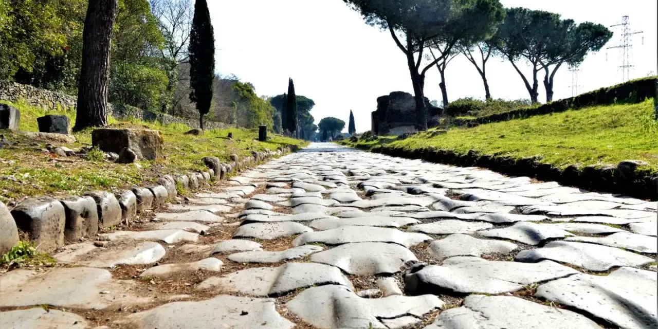 Ancient Roman cobblestone road lined with trees and ruins, leading towards the horizon.