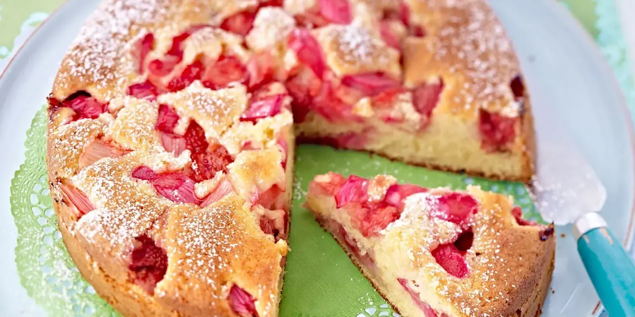 Ein saftiger Erdbeer Rhabarber Kuchen, bestäubt mit Puderzucker, wartet auf seinen Anschnitt. Ein Stück ist bereits auf einem grünen Tupfer platziert.