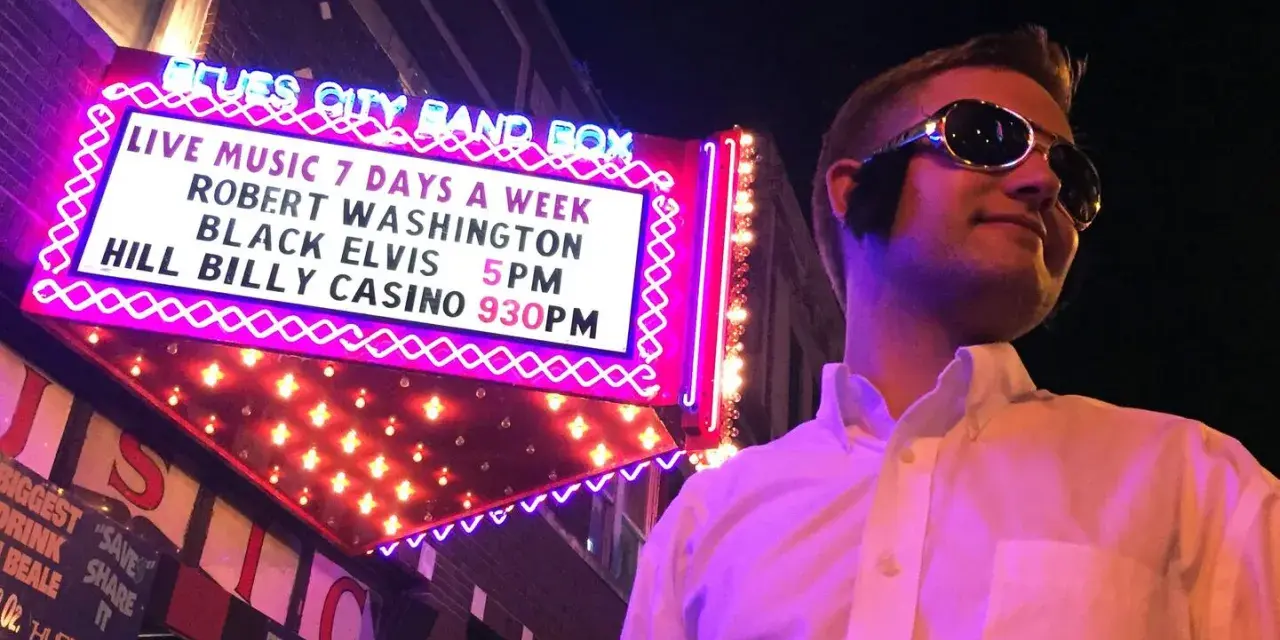 Mann im Elvis-Stil mit Sonnenbrille vor der "Blues City Band Box"-Neonreklame, die "Black Elvis" ankündigt.