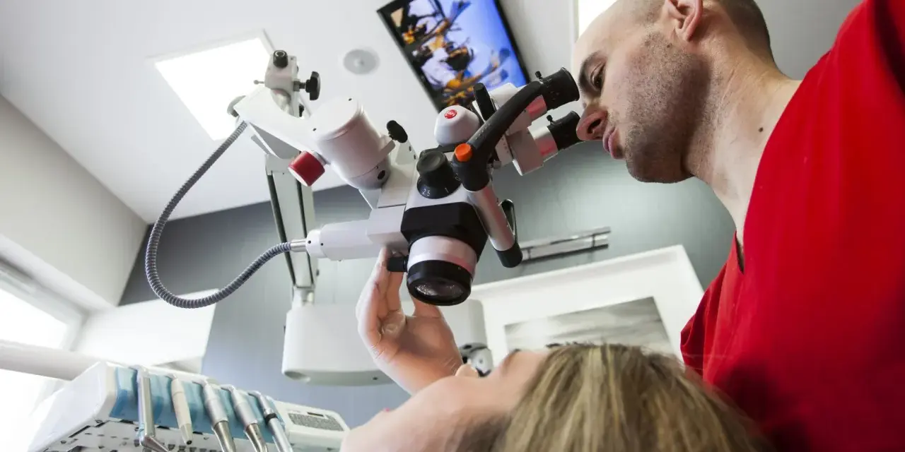 Dentysta w czerwonej bluzie bada pacjentkę przez mikroskop stomatologiczny.