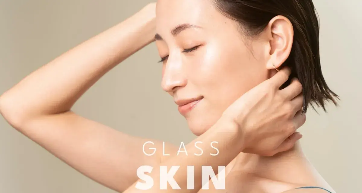 Kobieta z idealną, lśniącą cerą typu "glass skin", z dłońmi przy szyi i ramieniu.