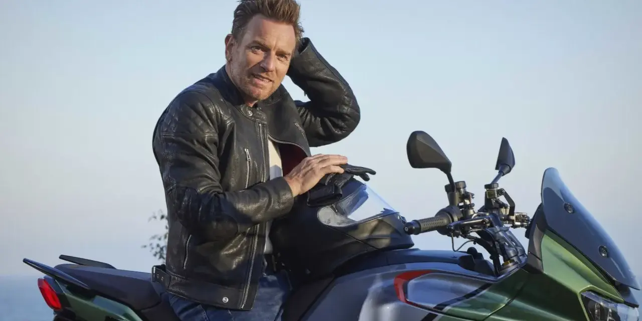 Ewan McGregor, con giacca di pelle, posa su una Moto Guzzi verde e grigia, pronto per un'avventura.