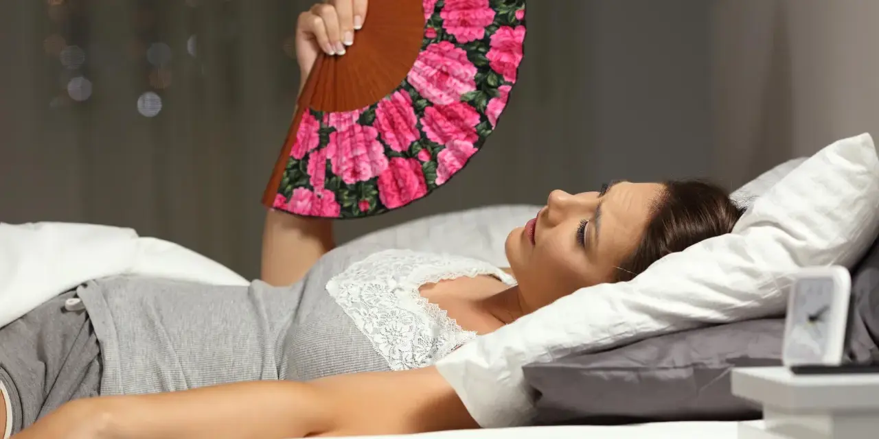 Frau liegt im Bett, nass geschwitzt, fächelt sich mit einem Blumenfächer Luft zu. Die Nacht ist heiß.