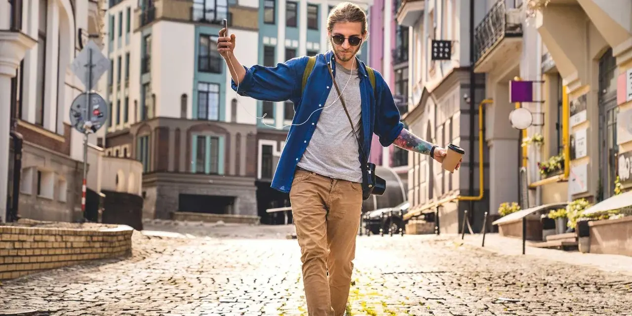 Giovane con caffè e smartphone cammina in città su un selciato, godendosi la musica e il paesaggio urbano.