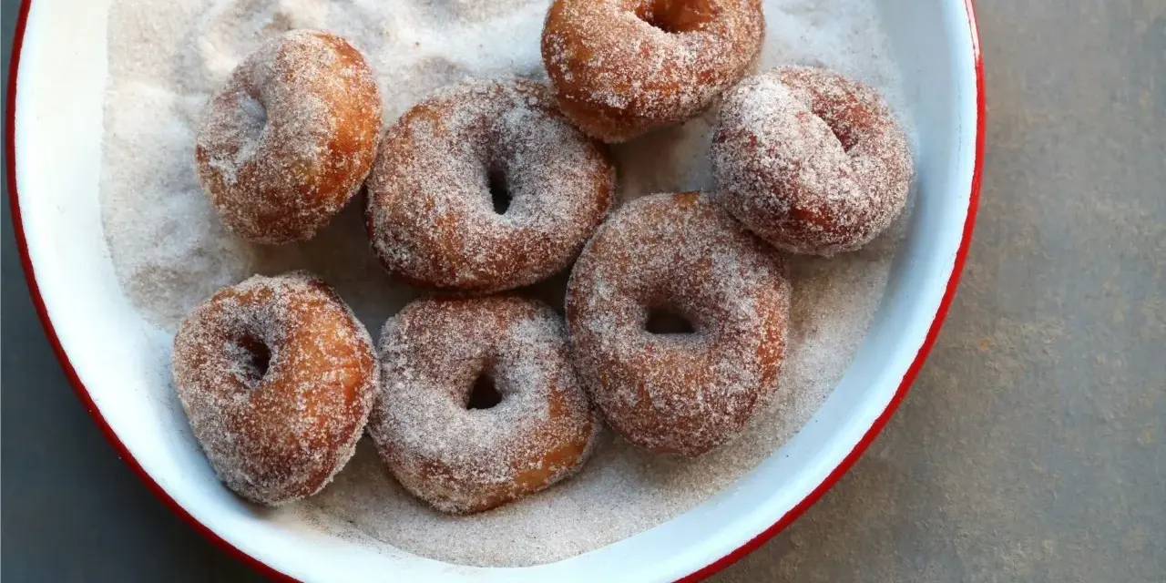 Små, nybakade donuts som rullats i socker och kanel ligger i en vit skål med röd kant.