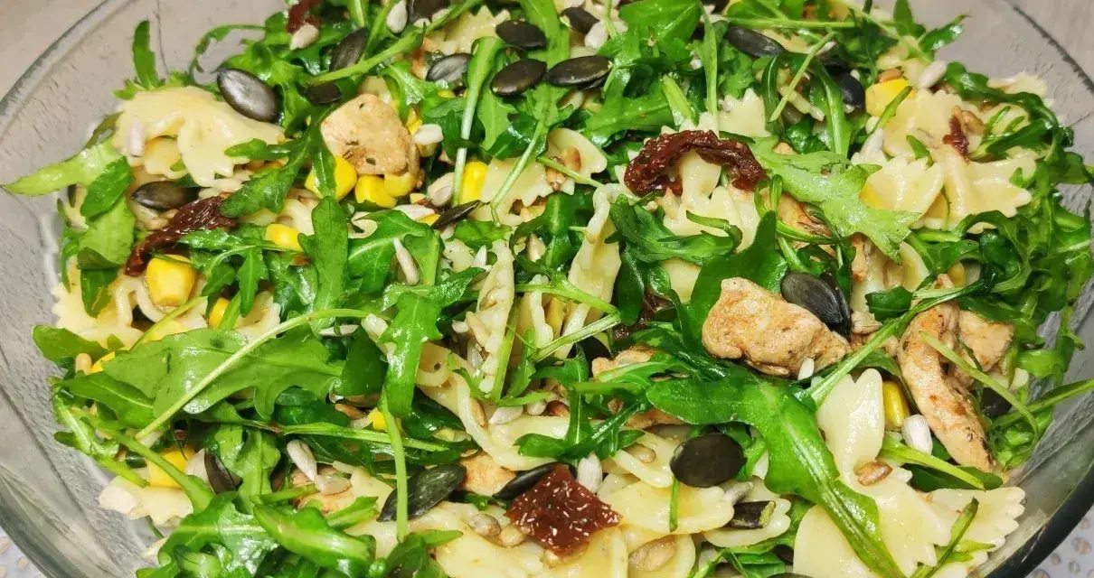 Kolorowa sałatka z makaronem farfalle, rukolą, kurczakiem, kukurydzą i suszonymi pomidorami, posypana pestkami dyni i słonecznika.