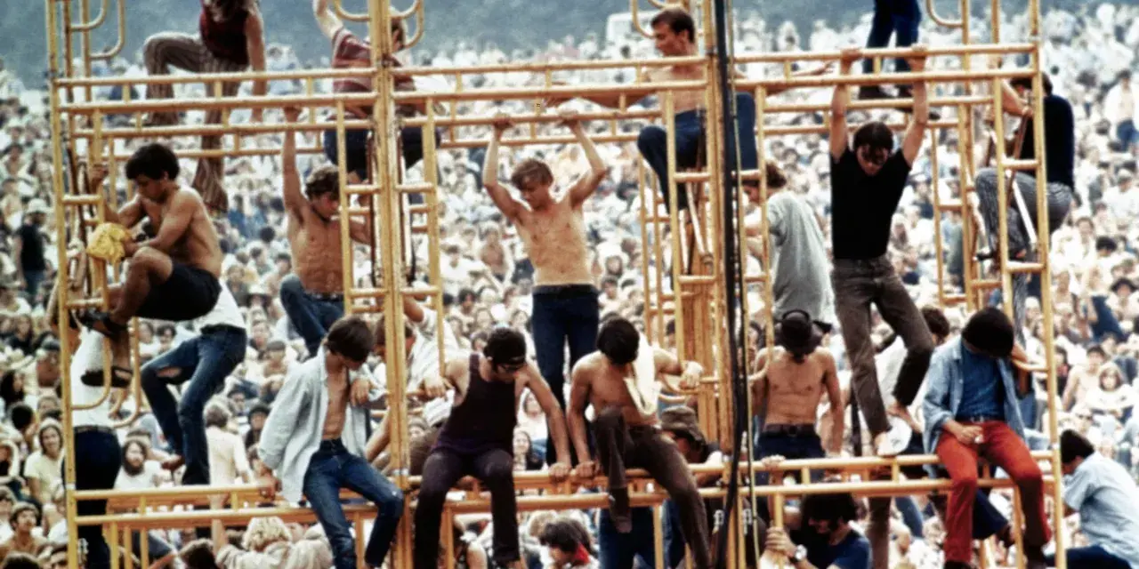 Tłum ludzi na festiwalu, niektórzy wspinają się na konstrukcję. To właśnie woodstock co to za wydarzenie?