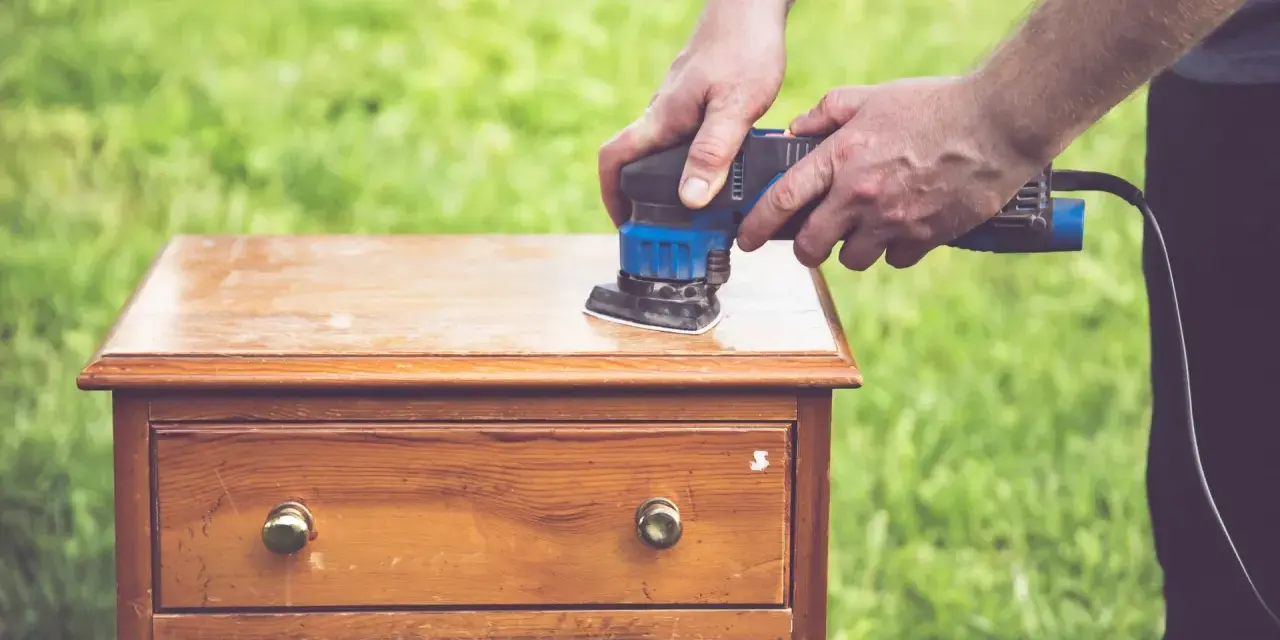 Un homme utilise une ponceuse pour le décapage mécanique d'une commode en bois, préparant le meuble pour une nouvelle finition.