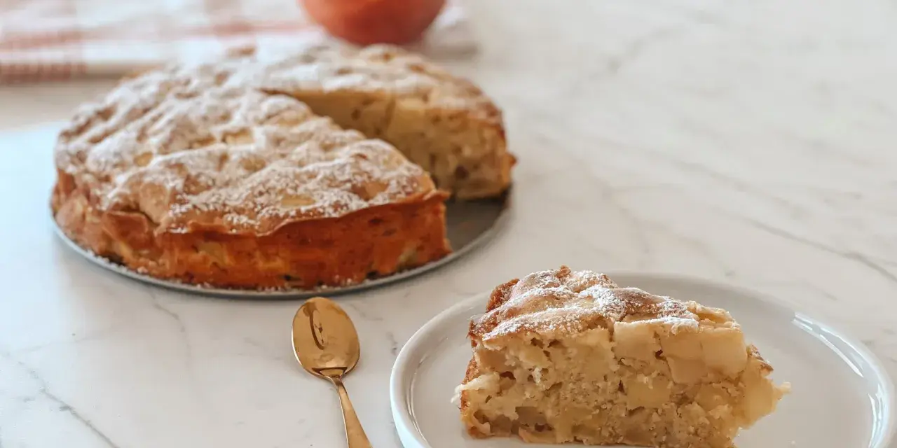 Une part de **crépiau aux pommes** saupoudrée de sucre glace, à côté d'un gâteau entier et d'une pomme. Une cuillère dorée est posée sur l'assiette.