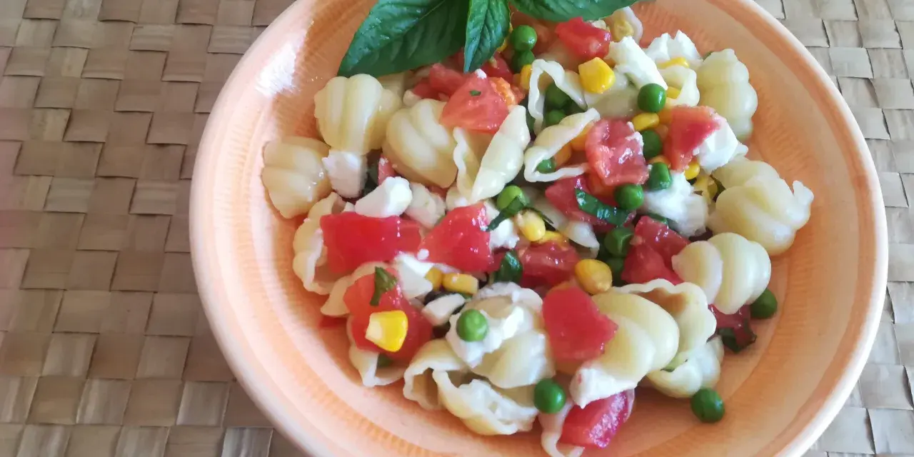 Un'appetitosa insalata di pasta vegetariana con pomodorini, piselli, mais e mozzarella, guarnita con basilico fresco.