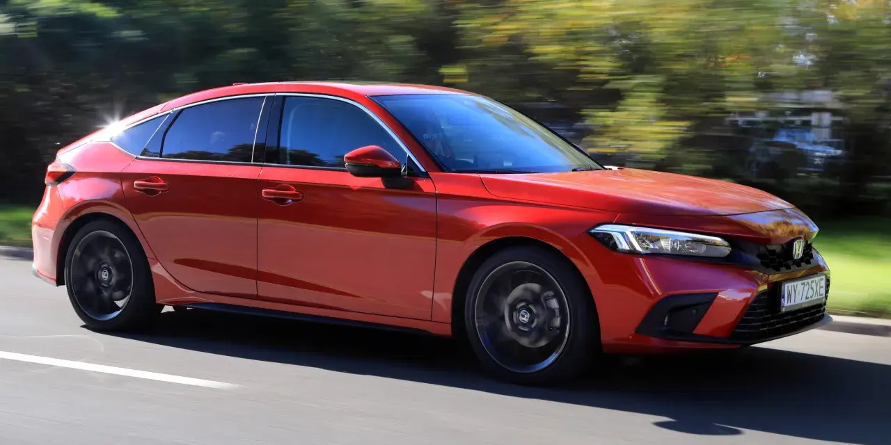 Czerwony Honda Civic 11. generacji pędzi po drodze, otoczony zielenią drzew.