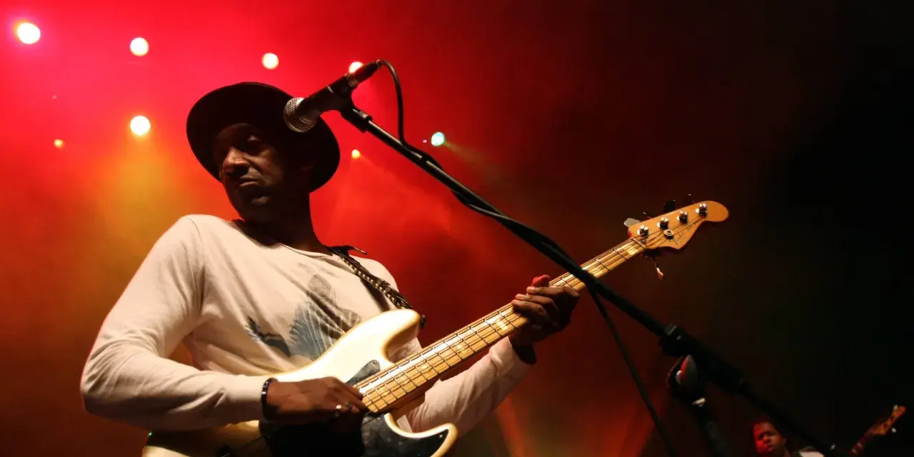Marcus Miller w czarnym kapeluszu gra na białej gitarze basowej na tle czerwonych świateł scenicznych.