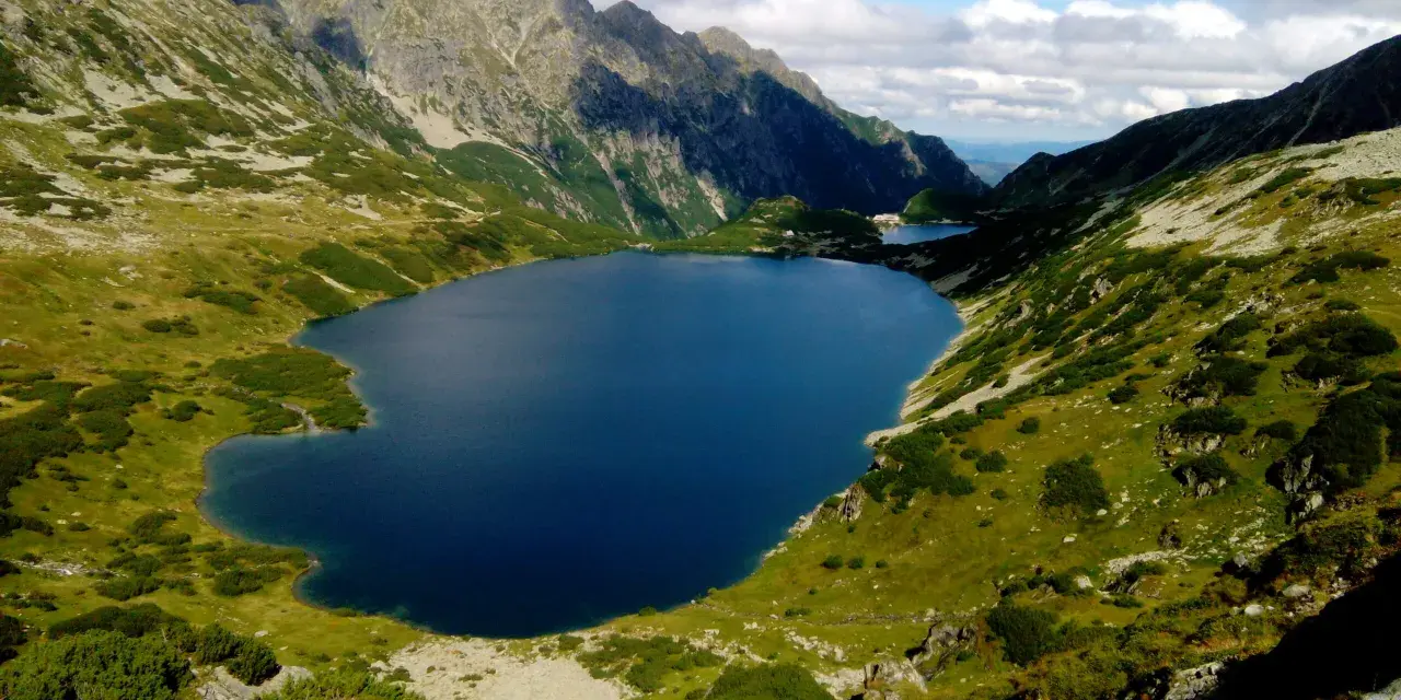 Malowniczy widok na Dolinę 5 Stawów. Głęboki, granatowy staw otoczony zielonymi zboczami gór i skalistymi szczytami.