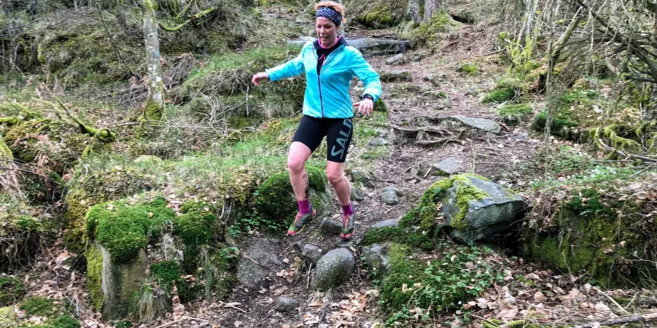 Kvinna springer i skogen, fokuserad på att springa marathon. Mossbeklädda stenar och träd skapar en utmanande terräng.
