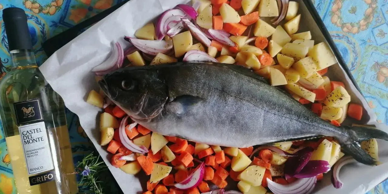 Ricciola al forno pronta per essere infornata con patate, carote e cipolle rosse. Accanto, una bottiglia di vino bianco.