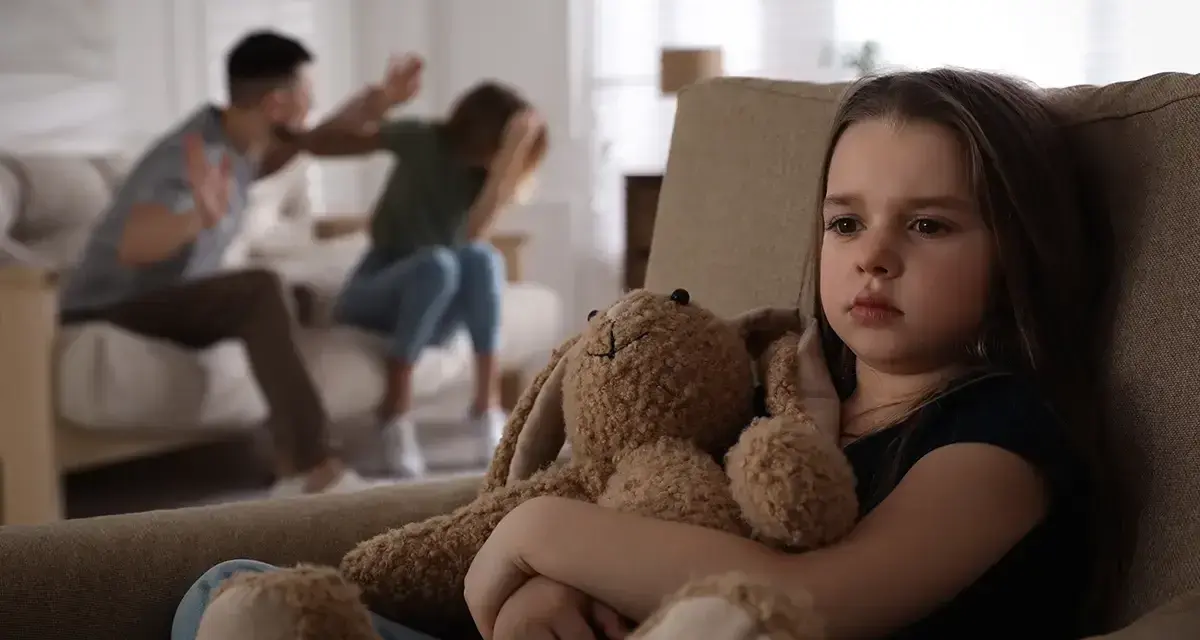 Ein trauriges Mädchen hält seinen Teddybär, während im Hintergrund Eltern streiten. Eine blitzscheidung ohne trennungsjahr hinterlässt Spuren.