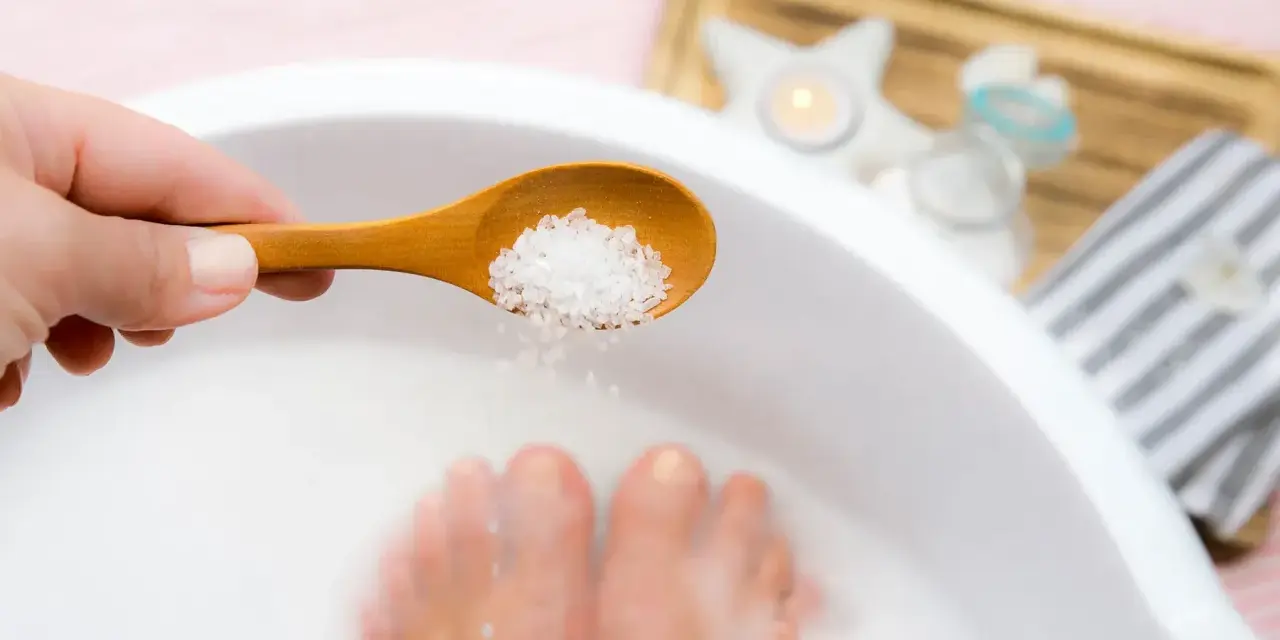 A hand adds epsom salt and baking soda to a foot soak, with feet submerged in the milky water.