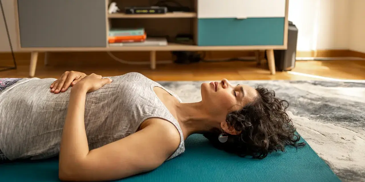 Frau liegt entspannt auf einer Matte, Hände auf dem Bauch, Augen geschlossen. Sie praktiziert progressive Muskelentspannung im Bett.