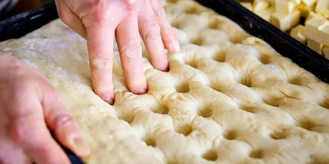 Hände drücken Vertiefungen in den Teig für ein Butterkuchen Rezept. Würfel aus Butter liegen bereit.