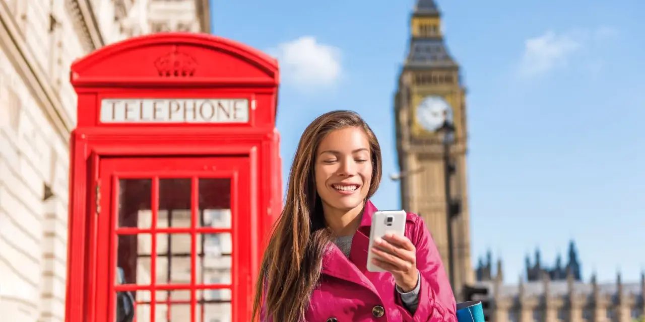 Uśmiechnięta kobieta z telefonem przed czerwoną budką telefoniczną i Big Benem. Pyta, czy w UK jest roaming, bo chce zadzwonić do domu.